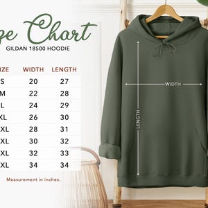 Puede incluir: Una sudadera con capucha Gildan 18500 verde oliva exhibida en una percha de madera. Una tabla de tallas muestra las medidas en pulgadas para las tallas S a 5XL. La tabla incluye medidas de ancho y largo para cada talla. La sudadera tiene un bolsillo delantero y una capucha con cord&oacute;n.