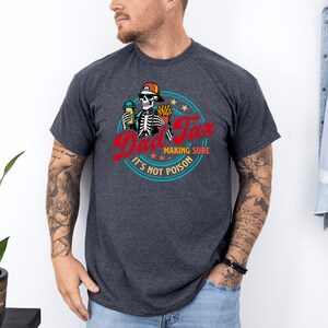 Pode incluir: Camiseta cinza escuro com um desenho de esqueleto usando um bon&eacute;, segurando uma bebida e batatas fritas, com o texto "Dad Tax" em vermelho e "Making Sure It's Not Poison" em um design circular. Uma pe&ccedil;a de roupa casual.