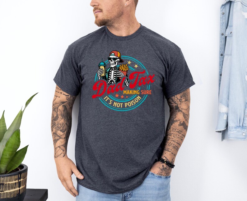 Pode incluir: Camiseta cinza escuro com um desenho de esqueleto usando um bon&eacute;, segurando uma bebida e batatas fritas, com o texto "Dad Tax" em vermelho e "Making Sure It's Not Poison" em um design circular. Uma pe&ccedil;a de roupa casual.