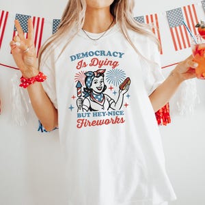 May include: White t-shirt with a retro graphic that reads "Democracy Is Dying But Hey-Nice Fireworks." The design features a woman holding a firework and a hot dog. American flags and red, white, and blue decorations are in the background.