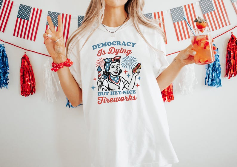 May include: White t-shirt with a retro graphic that reads "Democracy Is Dying But Hey-Nice Fireworks." The design features a woman holding a firework and a hot dog. American flags and red, white, and blue decorations are in the background.