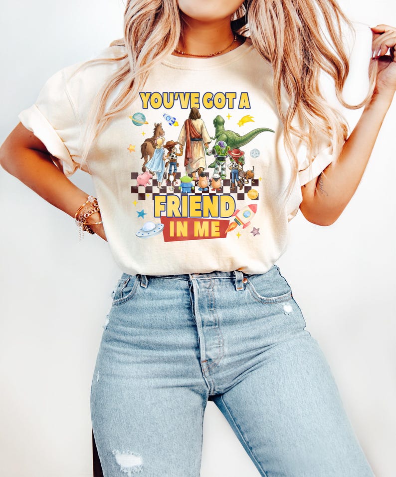 May include: A cream-colored short-sleeve t-shirt featuring a graphic print of a central figure walking with various cartoon characters on a checkered floor. The text reads YOU'VE GOT A FRIEND IN ME, surrounded by colorful space-themed icons and stars.