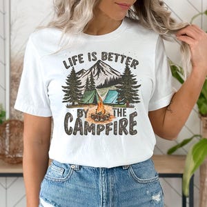 May include: White t-shirt with a graphic design of a campfire scene with mountains, trees, and tents. The text "Life is better by the campfire" is printed on the shirt.