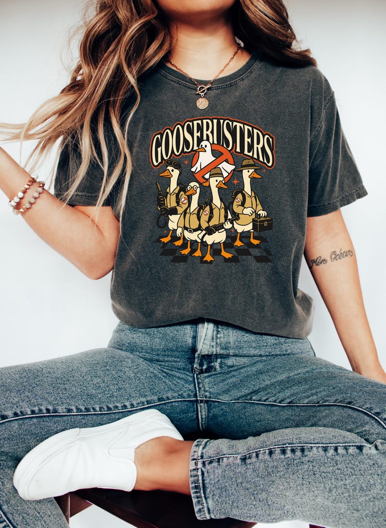 May include: Dark gray t-shirt with the text "GOOSEBUSTERS" in orange and white. The graphic features cartoon geese dressed as Ghostbusters. The geese are wearing khaki uniforms and carrying equipment. The shirt is a comfortable, casual style.