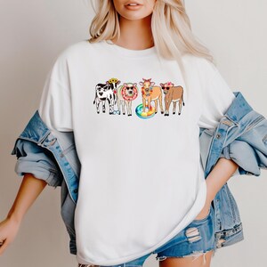Pu&ograve; includere: T-shirt bianco con una grafica colorata di cinque mucche dei cartoni animati. Ogni mucca indossa occhiali da sole, fasce floreali e salvagenti, creando un'atmosfera estiva e divertente. Il design &egrave; giocoso e accattivante.