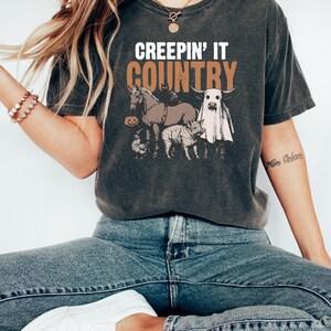 May include: Dark gray t-shirt with the text "CREEPIN' IT COUNTRY" in white and brown. The graphic features a horse, cat, cow, pig, chicken, and pumpkin. The shirt is worn with blue jeans and white shoes.