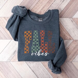 May include: A dark grey sweatshirt featuring the words "FALL vibes" in a checkered pattern of orange, green, and brown. The garment is folded on a light brown and white wooden surface.
