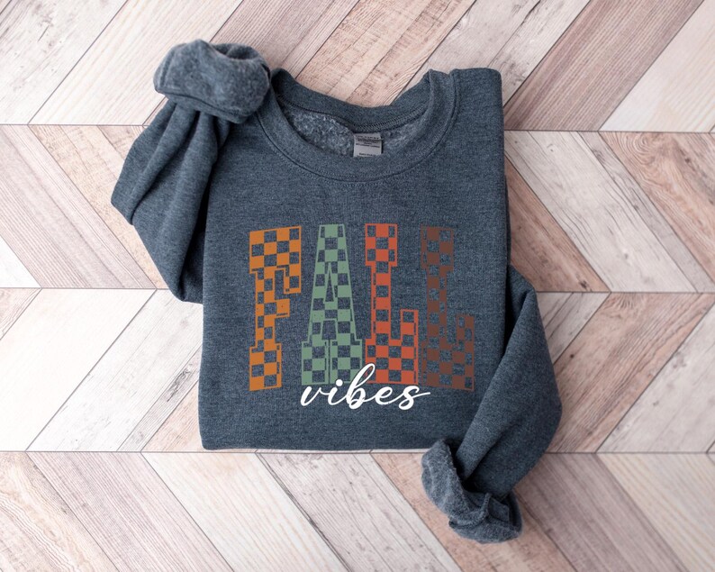 May include: A dark grey sweatshirt featuring the words "FALL vibes" in a checkered pattern of orange, green, and brown. The garment is folded on a light brown and white wooden surface.
