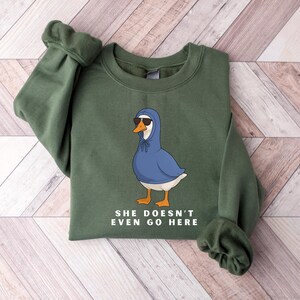 May include: An olive green sweatshirt with a cartoon goose wearing a blue hoodie and sunglasses. The goose has orange feet and a white chest. The text "SHE DOESN'T EVEN GO HERE" is printed below the goose.