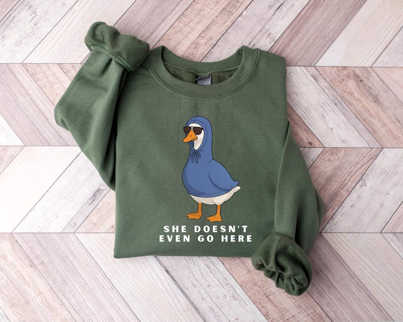May include: An olive green sweatshirt with a cartoon goose wearing a blue hoodie and sunglasses. The goose has orange feet and a white chest. The text "SHE DOESN'T EVEN GO HERE" is printed below the goose.