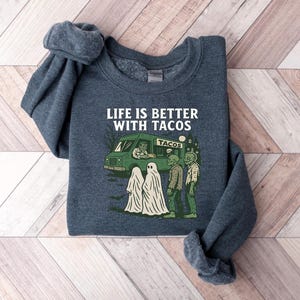 May include: A dark blue sweatshirt with the phrase "LIFE IS BETTER WITH TACOS" in white. The graphic features a taco truck, ghosts, and zombies. The sweatshirt is folded on a wooden surface.