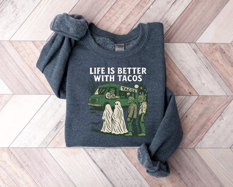 May include: A dark blue sweatshirt with the phrase "LIFE IS BETTER WITH TACOS" in white. The graphic features a taco truck, ghosts, and zombies. The sweatshirt is folded on a wooden surface.