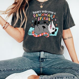 May include: Dark gray t-shirt with the text "Today's yoga pose is DOWNWARD SPIRAL" above a cartoon opossum in a yoga pose. The opossum is gray with a pink and orange tail, lying on a blue yoga mat. The shirt is worn with blue jeans and white shoes.