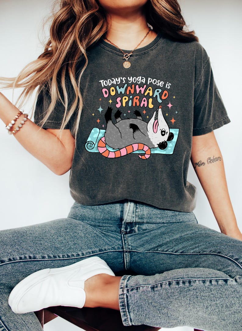 May include: Dark gray t-shirt with the text "Today's yoga pose is DOWNWARD SPIRAL" above a cartoon opossum in a yoga pose. The opossum is gray with a pink and orange tail, lying on a blue yoga mat. The shirt is worn with blue jeans and white shoes.