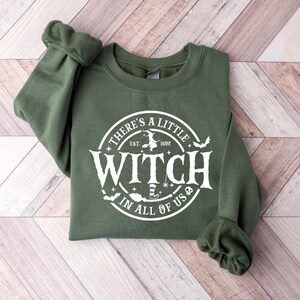 Op de afbeelding: Een olijfgroene sweatshirt met de witte tekst "THERE'S A LITTLE WITCH IN ALL OF US" in een cirkelvormig ontwerp. Het ontwerp bevat een heksensilhouet, vleermuizen en het jaar 1692. De sweatshirt ligt op een lichtbruin en wit houten oppervlak.