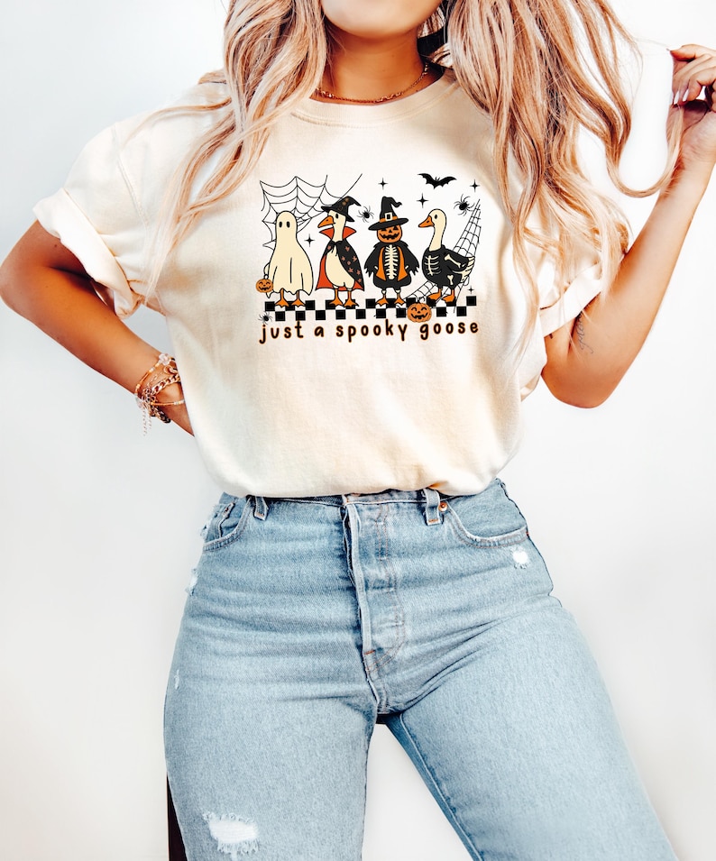 May include: Cream-colored t-shirt featuring a Halloween-themed graphic. The design includes four cartoon characters dressed as a ghost, vampire, scarecrow, and a goose, with the text "just a spooky goose". The shirt is paired with light blue jeans.