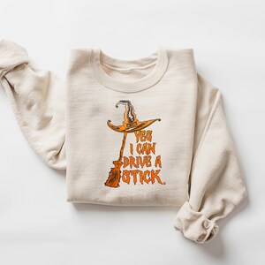 May include: A cream-colored sweatshirt with a Halloween-themed graphic. The graphic features a witch's hat and broom with the text "YES I CAN DRIVE A STICK" in orange. The sweatshirt is folded.
