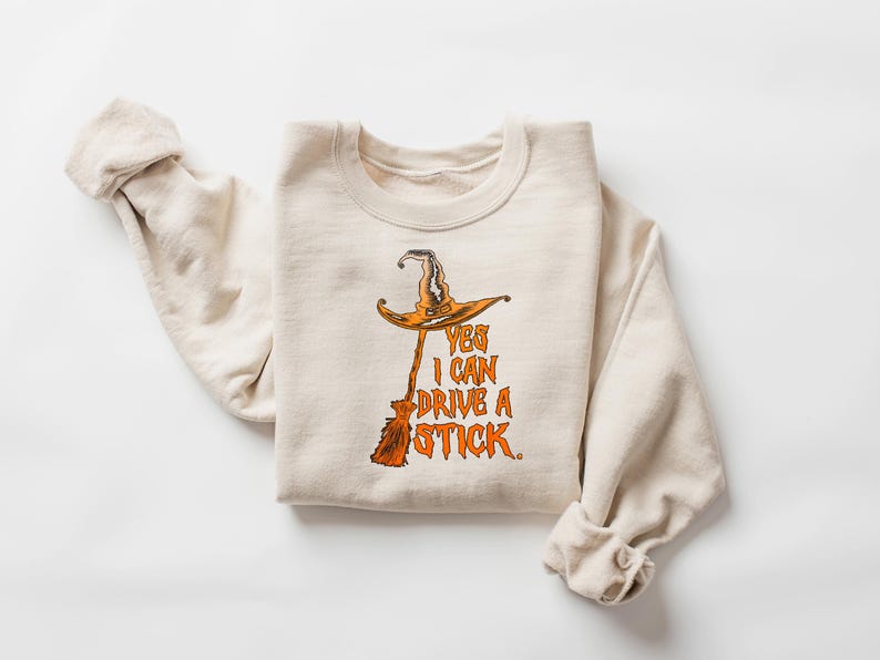 May include: A cream-colored sweatshirt with a Halloween-themed graphic. The graphic features a witch's hat and broom with the text "YES I CAN DRIVE A STICK" in orange. The sweatshirt is folded.