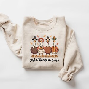 Pu&ograve; includere: Felpa beige con una grafica di quattro oche dei cartoni animati vestite per il Ringraziamento. Le oche indossano cappelli, grembiuli e camicie a quadri. Il testo "just a thankful goose" &egrave; sotto.