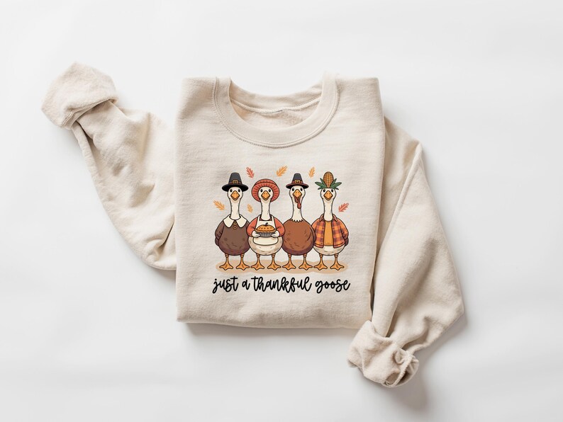 Pu&ograve; includere: Felpa beige con una grafica di quattro oche dei cartoni animati vestite per il Ringraziamento. Le oche indossano cappelli, grembiuli e camicie a quadri. Il testo "just a thankful goose" &egrave; sotto.