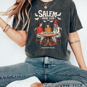 Puede incluir: Camiseta gris oscuro con un gr&aacute;fico de tres figuras alrededor de una mesa, con el texto "Salem Book Club - 1692" y murci&eacute;lagos. La camiseta tambi&eacute;n incluye el texto "Massachusetts Est. 1692".