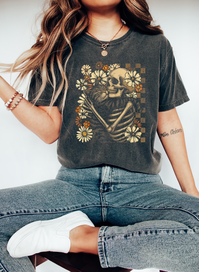 May include: Dark gray t-shirt featuring a skeleton embracing a cat, surrounded by flowers. The design includes a checkered background. The shirt is worn with blue jeans and white slip-on shoes. The overall theme is a blend of gothic and floral elements.