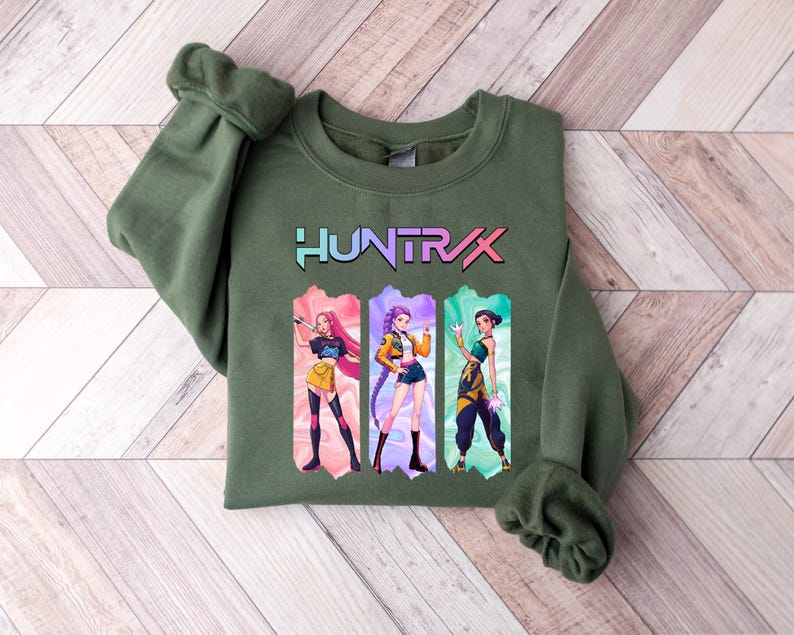 May include: Olive green sweatshirt with the word "HUNTRAX" in a colorful font. Below the text are three anime-style illustrations of women in action poses, each in a different outfit. The sweatshirt has a crew neck and long sleeves.