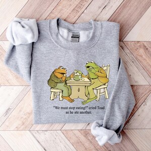 Puede incluir: Una sudadera gris jaspeada con una ilustraci&oacute;n de dos ranas en una mesa. Las ranas visten ropa y comen de un cuenco. El texto "We must stop eating!" cried Toad as he ate another est&aacute; impreso debajo.