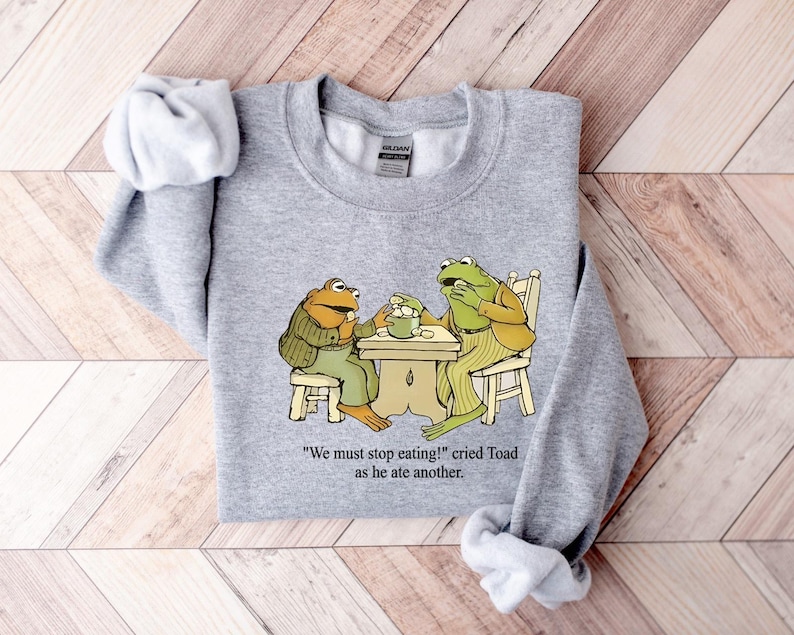 Puede incluir: Una sudadera gris jaspeada con una ilustraci&oacute;n de dos ranas en una mesa. Las ranas visten ropa y comen de un cuenco. El texto "We must stop eating!" cried Toad as he ate another est&aacute; impreso debajo.