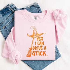 May include: A light pink sweatshirt with the text "YES I CAN DRIVE A STICK" in orange. The graphic features a witch's hat and broom. The sweatshirt is laid on top of blue jeans and a white surface.