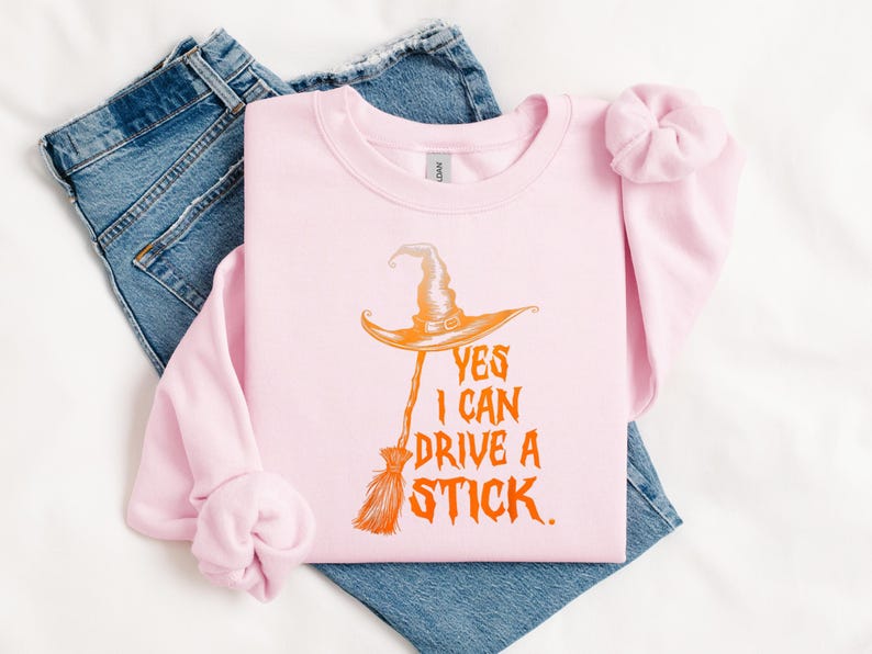 May include: A light pink sweatshirt with the text "YES I CAN DRIVE A STICK" in orange. The graphic features a witch's hat and broom. The sweatshirt is laid on top of blue jeans and a white surface.