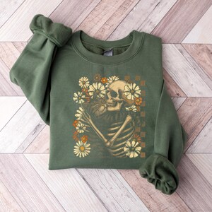 May include: An olive green sweatshirt featuring a graphic of a skeleton embracing a bouquet of flowers. The skeleton is cream-colored, and the flowers are in shades of cream, orange, and brown. The design is set against a checkered background.