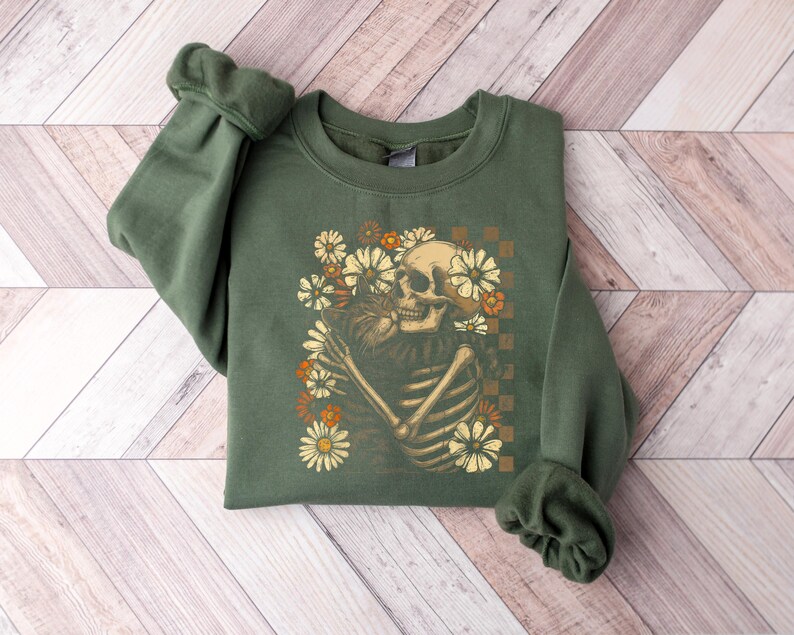 May include: An olive green sweatshirt featuring a graphic of a skeleton embracing a bouquet of flowers. The skeleton is cream-colored, and the flowers are in shades of cream, orange, and brown. The design is set against a checkered background.