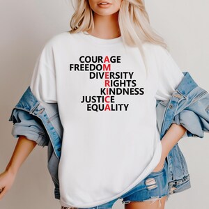 Puede incluir: Camiseta blanca con la palabra "AMERICA" escrita verticalmente con las palabras "Coraje, Libertad, Diversidad, Derechos, Amabilidad, Justicia, Igualdad". Las letras "A", "R", "I" y "A" son rojas.