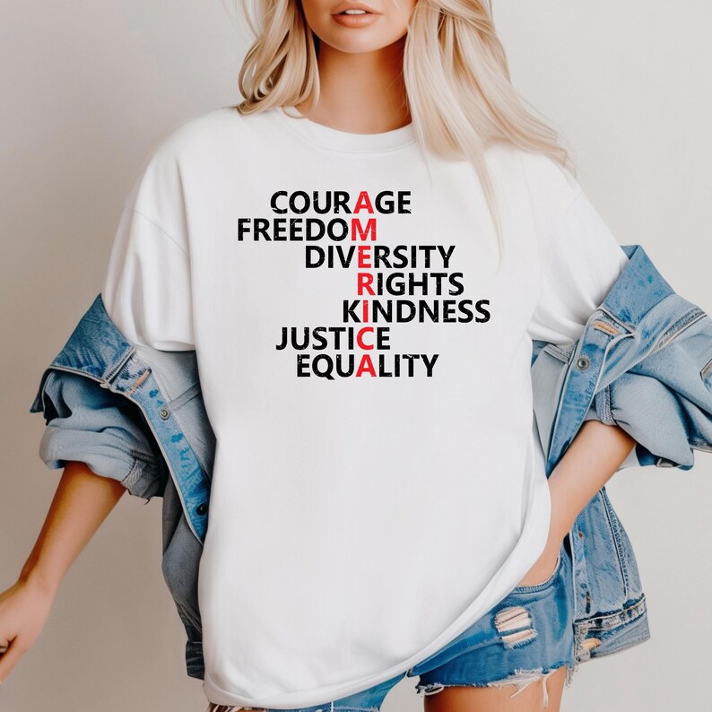 Puede incluir: Camiseta blanca con la palabra "AMERICA" escrita verticalmente con las palabras "Coraje, Libertad, Diversidad, Derechos, Amabilidad, Justicia, Igualdad". Las letras "A", "R", "I" y "A" son rojas.