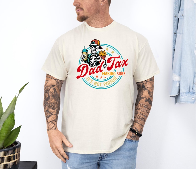 Pode incluir: Camiseta creme com um gr&aacute;fico circular. O gr&aacute;fico apresenta o texto "Dad Tax" em vermelho, um esqueleto usando chap&eacute;u e &oacute;culos de sol, segurando uma bebida e batatas fritas, e a frase "Making Sure It's Not Poison".