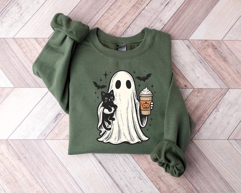 May include: Green sweatshirt with a Halloween design. The graphic shows a ghost holding a black cat and a pumpkin spice latte. The design also includes two bats and stars. The sweatshirt has long sleeves and a crew neck.