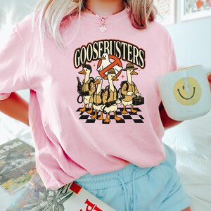 May include: Pink t-shirt featuring a graphic of five cartoon geese dressed as Ghostbusters, with the text "GOOSEBUSTERS" above them. The geese stand on a black and white checkered pattern.