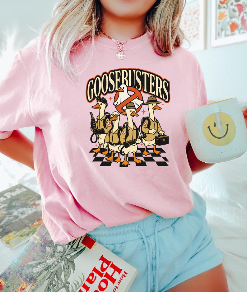 May include: Pink t-shirt featuring a graphic of five cartoon geese dressed as Ghostbusters, with the text "GOOSEBUSTERS" above them. The geese stand on a black and white checkered pattern.