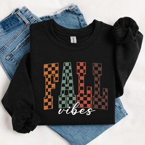 May include: Black sweatshirt with the words "FALL vibes" in a checkered pattern. The word "FALL" is in orange, green, and brown checkered letters. The word "vibes" is written in a cursive font in white. The sweatshirt is on top of blue jeans.