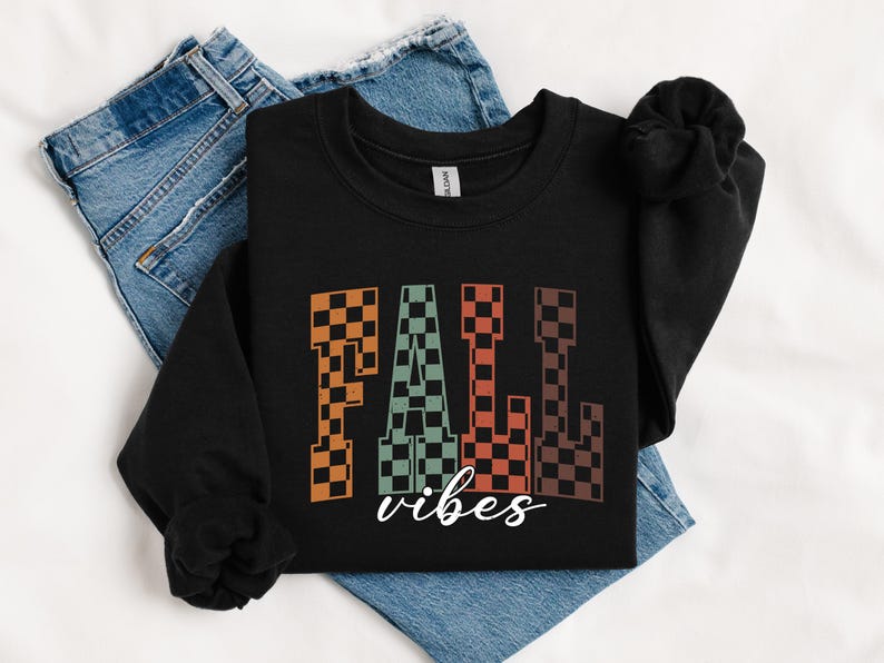 May include: Black sweatshirt with the words "FALL vibes" in a checkered pattern. The word "FALL" is in orange, green, and brown checkered letters. The word "vibes" is written in a cursive font in white. The sweatshirt is on top of blue jeans.