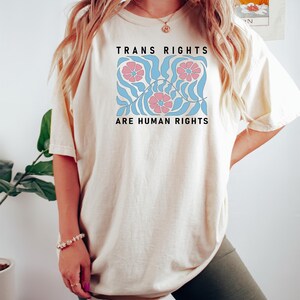 May include: A person wearing a light beige t-shirt with the text "Trans Rights Are Human Rights" and a floral design in pink, blue, and white. The person is also wearing dark green cycling shorts.