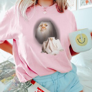 Puede incluir: Camiseta rosa claro con un dise&ntilde;o de gato fantasma. El gr&aacute;fico muestra la cara de un gato asomando por una s&aacute;bana blanca, con un segundo gato con disfraz de fantasma debajo. Una taza blanca con una carita sonriente amarilla se sostiene en la mano.