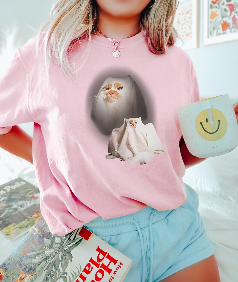 Puede incluir: Camiseta rosa claro con un dise&ntilde;o de gato fantasma. El gr&aacute;fico muestra la cara de un gato asomando por una s&aacute;bana blanca, con un segundo gato con disfraz de fantasma debajo. Una taza blanca con una carita sonriente amarilla se sostiene en la mano.