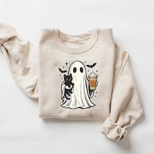 May include: Beige sweatshirt with a Halloween graphic. The design shows a ghost holding a black cat and a pumpkin spice latte. Bats and stars are also included in the illustration. A cosy and festive apparel item.