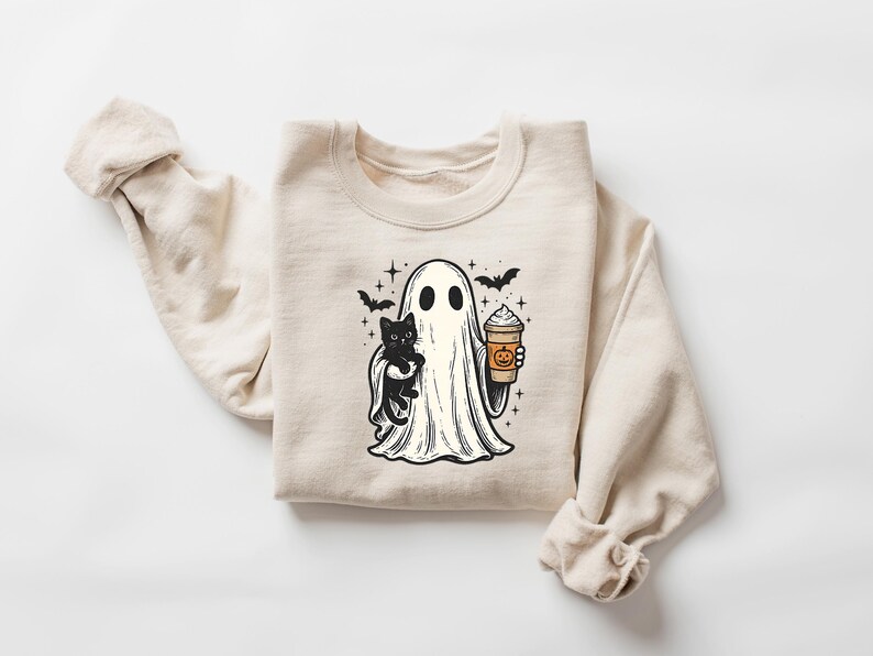 May include: Beige sweatshirt with a Halloween graphic. The design shows a ghost holding a black cat and a pumpkin spice latte. Bats and stars are also included in the illustration. A cosy and festive apparel item.