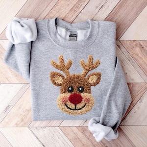 May include: A light gray sweatshirt featuring a reindeer design. The reindeer has brown antlers, a brown face, a red nose, and a smiling mouth. The sweatshirt is folded on a wooden surface.