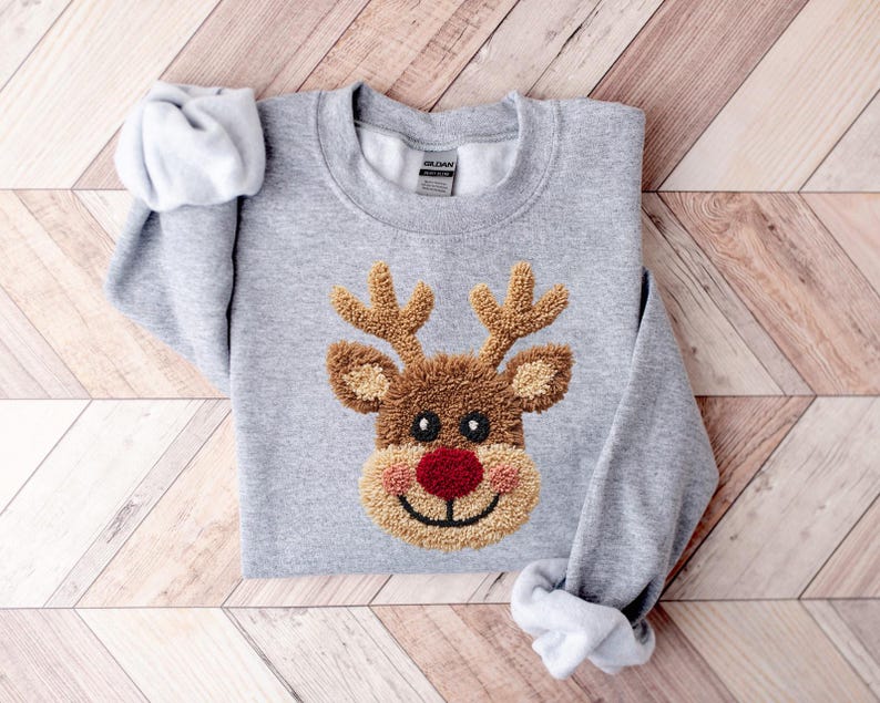 May include: A light gray sweatshirt featuring a reindeer design. The reindeer has brown antlers, a brown face, a red nose, and a smiling mouth. The sweatshirt is folded on a wooden surface.