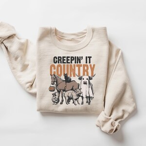 May include: A cream-colored sweatshirt with the text "CREEPIN' IT COUNTRY" in black and brown. The graphic features a horse, cat, cow, pig, and chicken, all with Halloween-themed costumes. The sweatshirt is made of a soft material.