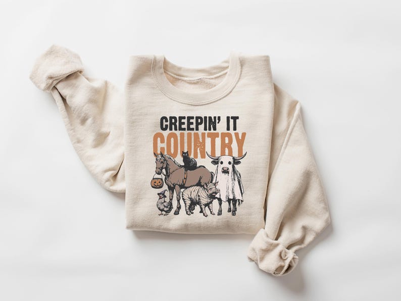 May include: A cream-colored sweatshirt with the text "CREEPIN' IT COUNTRY" in black and brown. The graphic features a horse, cat, cow, pig, and chicken, all with Halloween-themed costumes. The sweatshirt is made of a soft material.